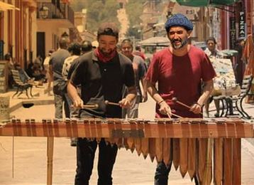  due uomini suonano una marimba in una strada affollata. Sullo sfondo si vedono persone e negozi colorati.