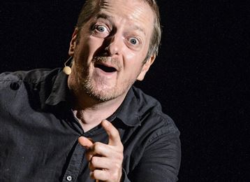 A cheerful man stands on stage and points his finger at the audience. He is wearing a black shirt and appears lively and entertaining.
