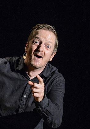 Un homme joyeux se tient sur scène et pointe du doigt le public. Il porte une chemise noire et a l'air vif et divertissant.