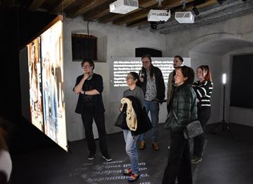 Un groupe de personnes se tient dans une salle d'exposition moderne. Sur le mur, de grands écrans de projection affichent des images et des textes.