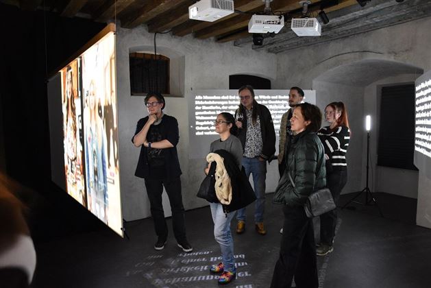 A group of people is standing in a modern exhibition space. Large projection surfaces with images and texts are hanging on the wall.
