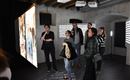 A group of people is standing in a modern exhibition space. Large projection surfaces with images and texts are hanging on the wall.