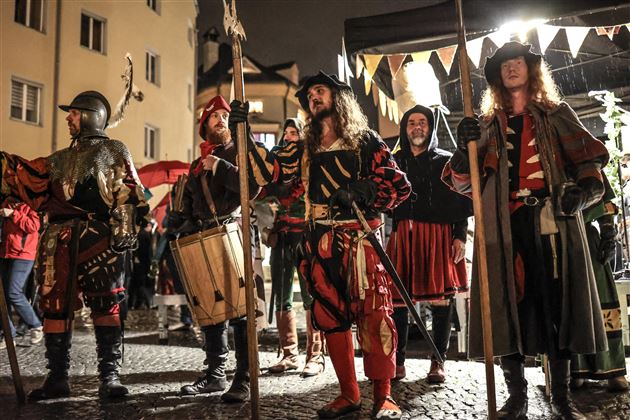 Un gruppo di uomini in costume storico si trova in un mercato festivo. Indossano diverse armature e acconciature e presentano un'atmosfera vivace.
