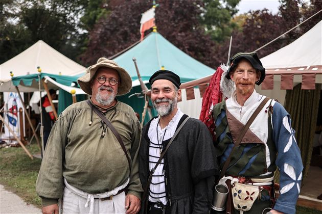 Tre uomini in costumi storici sorridevano davanti a tende colorate. Fanno parte di un festival medievale.
