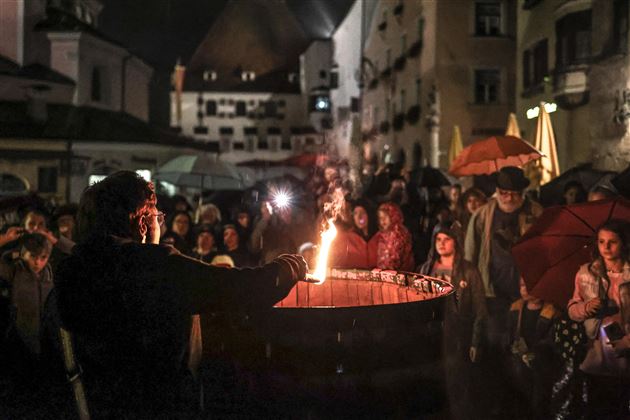 Una folla di persone sta sotto ombrelli e guarda un artista che fa un'esibizione con una torcia in mano. La scena si svolge di notte in una vecchia strada di città.