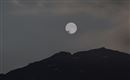 Ein großer, leuchtender Mond steht über einem dunklen Berg. Der Himmel ist leicht bewölkt und wirkt mystisch.