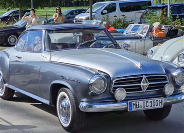 Ein silberner Oldtimer mit klassischem Design steht auf der Straße. Im Hintergrund sind Menschen und weitere Autos zu sehen.