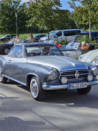 Ein silberner Oldtimer mit klassischem Design steht auf der Straße. Im Hintergrund sind Menschen und weitere Autos zu sehen.