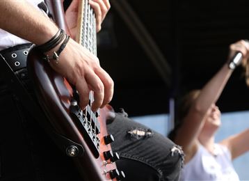 Eine Person spielt Gitarre auf einer Bühne. Im Hintergrund sieht man einen anderen Musiker beim Singen.