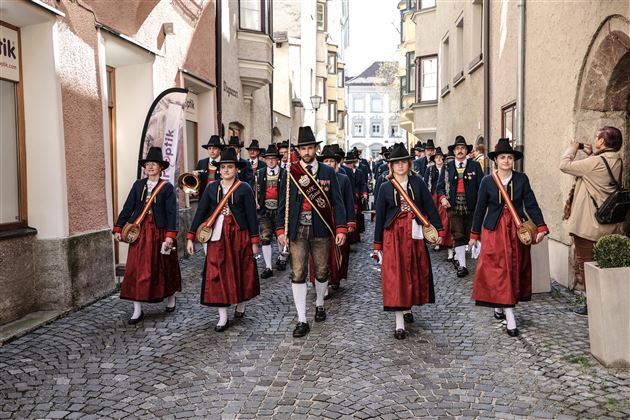 Eine Gruppe von Menschen in traditioneller Kleidung marschiert durch eine enge Straße. Sie tragen historische Trachten und Hüte und scheinen an einer Festlichkeit teilzunehmen.