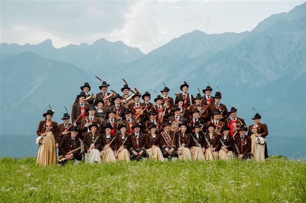 Un grande gruppo di persone in abiti tradizionali è in piedi su un prato davanti a un paesaggio montano. Tengono strumenti musicali e sorridono verso la fotocamera.