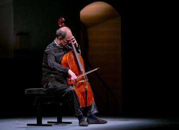 A cellist sits on a bench and plays his instrument. The stage is dimly lit, creating an intimate atmosphere.