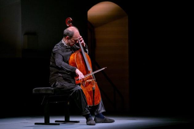 Ein Cellist sitzt auf einer Bank und spielt sein Instrument. Die Bühne ist schwach beleuchtet, was eine intime Atmosphäre schafft.