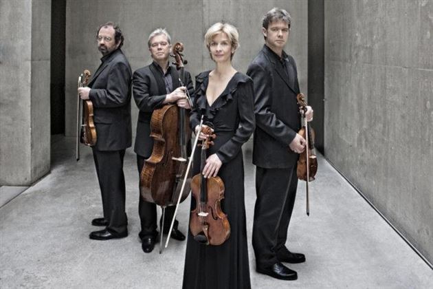 Eine professionelle Musikergruppe steht in eleganter Kleidung in einem minimalistischen Raum. Sie halten verschiedene Streichinstrumente und wirken aufmerksam.