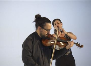 Un musicien joue du violon, tandis qu'une chanteuse chante au premier plan. Les deux sont représentés devant un fond neutre.