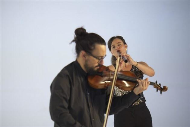 Ein Musiker spielt die Violine, während eine Sängerin im Vordergrund singt. Die beiden sind vor einem neutralen Hintergrund abgebildet.