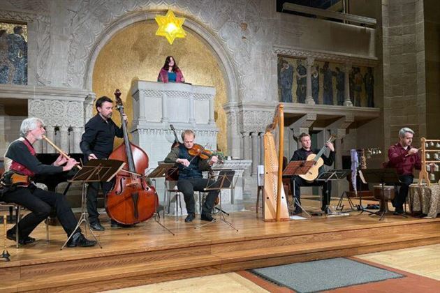 Eine musikalische Aufführung in einem festlichen Raum. Sechs Musiker spielen verschiedene Instrumente vor einer dekorativen Kulisse mit einer Statue.