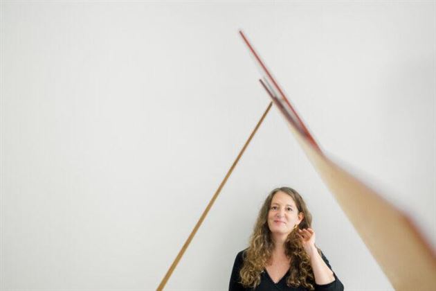 Eine Frau mit lockigem Haar steht vor einer weißen Wand. Sie lächelt und schaut nachdenklich auf einen schrägen Gegenstand, der ins Bild ragt.