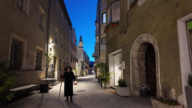 Eine schmale Gasse zur blauen Stunde mit historischen Gebäuden. Eine Person steht mit einer Laterne in der Mitte der Straße.