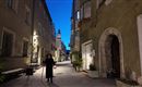 Eine schmale Gasse zur blauen Stunde mit historischen Gebäuden. Eine Person steht mit einer Laterne in der Mitte der Straße.