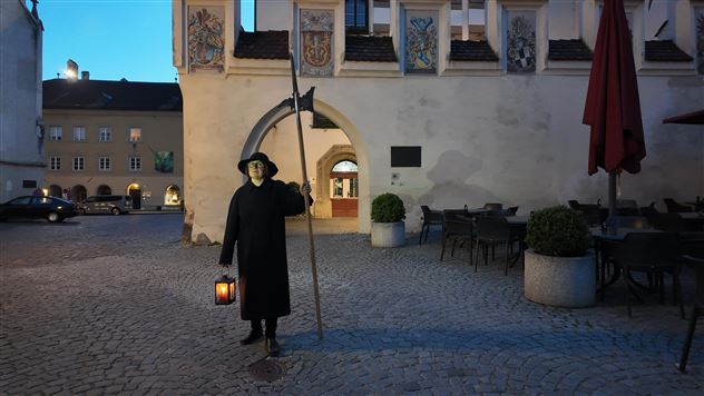 Eine Person in schwarzer Kleidung steht mit einer Laterne und einem Stock in einem historischen Platz. Die Szenerie ist abends, mit einem ruhigen, stimmungsvolle Licht.