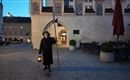 Eine Person in schwarzer Kleidung steht mit einer Laterne und einem Stock in einem historischen Platz. Die Szenerie ist abends, mit einem ruhigen, stimmungsvolle Licht.
