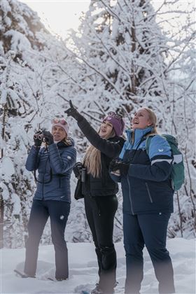 Drei Frauen stehen im Schnee und schauen nach oben. Sie lachen und wirken begeistert von der winterlichen Landschaft.
