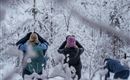 Drei Personen stehen im verschneiten Wald und schauen nach oben. Die Bäume sind mit Schnee bedeckt, und die Landschaft wirkt winterlich und friedlich.