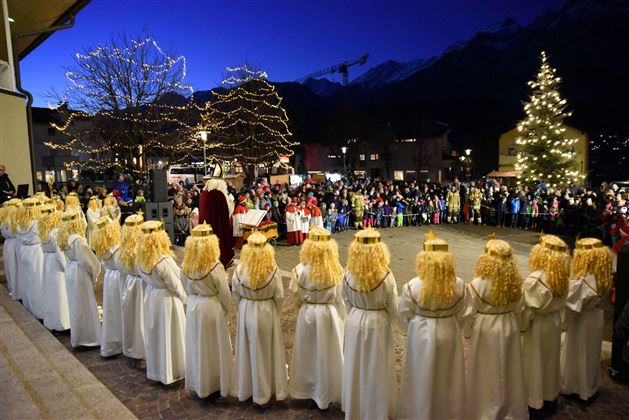 Una festosa assemblea con molti bambini in abiti bianchi e capelli biondi è in primo piano. Sullo sfondo si vede un albero di Natale e una grande folla.
