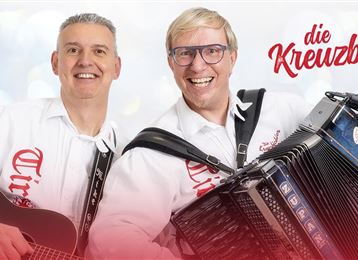 Zwei Musiker lächeln in die Kamera. Der eine spielt Gitarre und der andere ein Akkordeon.