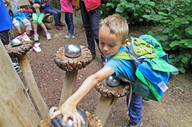 Een jongen speelt met een interactieve installatie in het bos. Op de achtergrond zijn andere kinderen te zien die ook betrokken zijn.