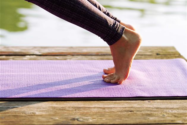Eine Person steht auf einem lilafarbenen Yoga-Matte am Rand eines Holzstegs. Im Hintergrund ist Wasser zu sehen.