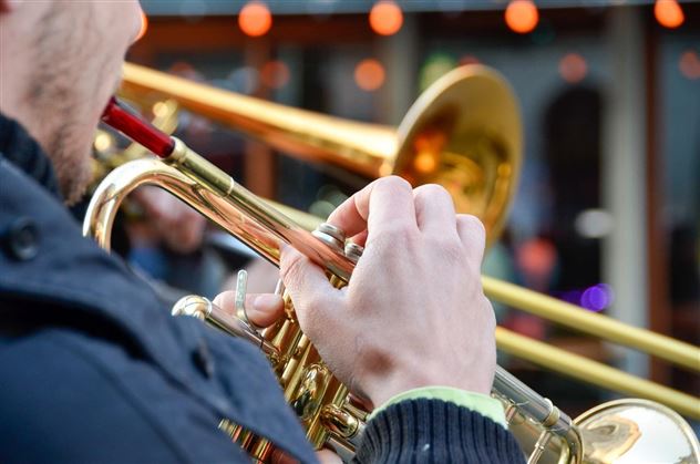 Ein Musiker spielt eine Trompete in einer lebhaften Umgebung. Seine Hand hält das Instrument, während im Hintergrund verschwommene Menschen sichtbar sind.