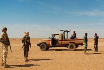 Un gruppo di soldati si trova in un desolato paesaggio desertico. Un veicolo blindato è visibile sullo sfondo.