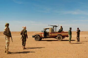 Een groep soldaten staat in een schrale woestijnlandschap. Een gepantserd voertuig is op de achtergrond te zien.