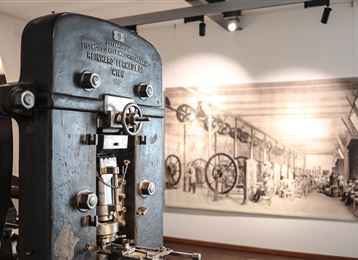 Eine alte Maschine in einem Museum, die die industrielle Geschichte symbolisiert. Im Hintergrund ist eine historische Zeichnung einer Fabrik zu sehen.