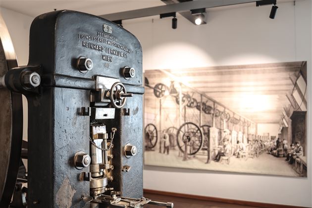 Eine alte Maschine in einem Museum, die die industrielle Geschichte symbolisiert. Im Hintergrund ist eine historische Zeichnung einer Fabrik zu sehen.