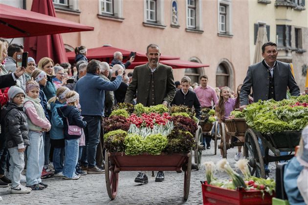 Eine belebte Straßenveranstaltung mit Menschen und Verkaufswagen voller frischer Blumen und Gemüse. Besucher beobachten die Szenen neugierig und freut sich über die festliche Atmosphäre.