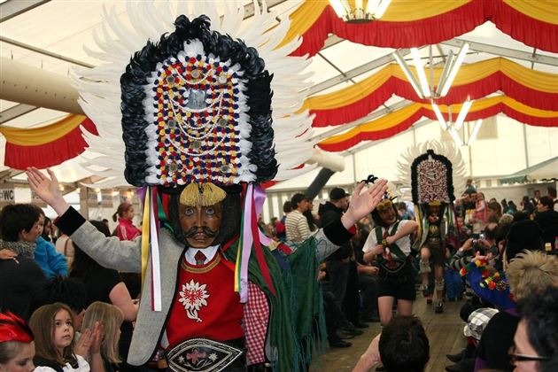 A festive parade with people in colorful traditional costumes and large hats. There is a lot going on, and the crowd is enthusiastically applauding.