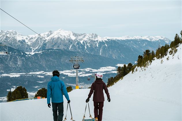 Ein paar Skifahrer gehen auf einer schneebedeckten Piste mit ihren Schlitten. Im Hintergrund sind beeindruckende Berge und ein Sessellift zu sehen.
