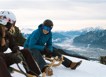 Deux personnes sont assises sur des traîneaux dans la neige et profitent de la vue sur le paysage montagneux. Les montagnes environnantes sont recouvertes de neige et le ciel est nuageux.