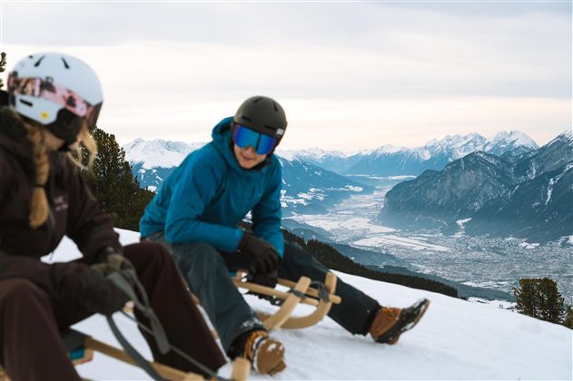 Zwei Personen sitzen auf Schlitten im Schnee und genießen die Aussicht auf die Berglandschaft. Die umliegenden Berge sind schneebedeckt und der Himmel ist bewölkt.