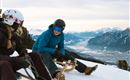 Zwei Personen sitzen auf Schlitten im Schnee und genießen die Aussicht auf die Berglandschaft. Die umliegenden Berge sind schneebedeckt und der Himmel ist bewölkt.