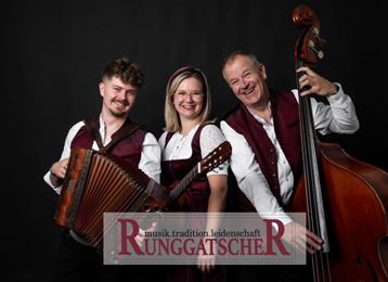 Eine Musikgruppe bestehend aus drei Mitgliedern, die lächelnd vor einem schwarzen Hintergrund posieren. Sie halten ein Akkordeon und einen Kontrabass und tragen traditionelle Kleidung.