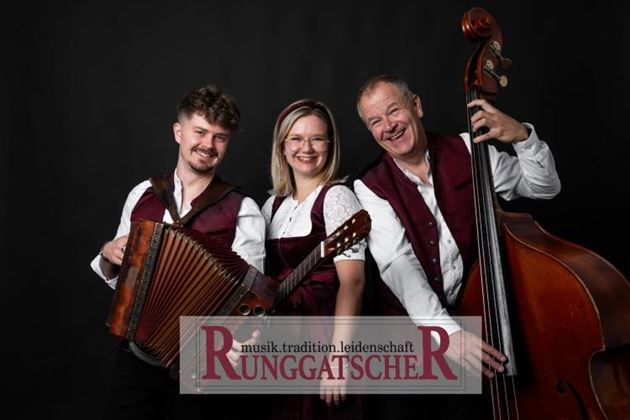 Eine Musikgruppe bestehend aus drei Mitgliedern, die lächelnd vor einem schwarzen Hintergrund posieren. Sie halten ein Akkordeon und einen Kontrabass und tragen traditionelle Kleidung.