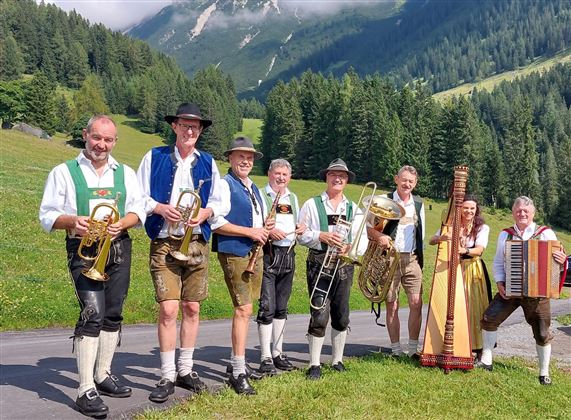 Eine Gruppe von Musikern in traditioneller Tracht steht in einer grünen Landschaft. Im Hintergrund sind Berge und Wälder zu sehen.
