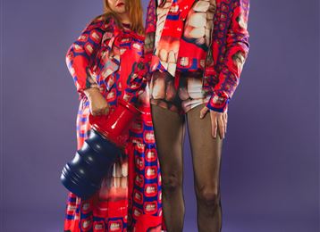 Deux mannequins dans des tenues colorées et saisissantes se tiennent devant un fond violet. L'un des mannequins tient un grand objet bleu dans la main.