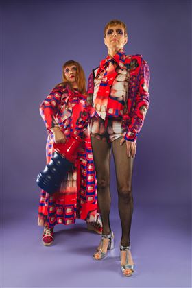 Zwei Models in auffälligen, bunten Outfits stehen vor einem lilafarbenen Hintergrund. Eines der Models hält einen großen, blauen Gegenstand in der Hand.