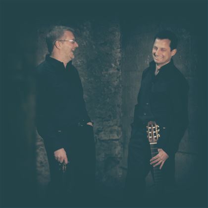 Deux hommes en vêtements noirs se tiennent côte à côte et sourient. L'un d'eux tient une guitare et ils semblent discuter.