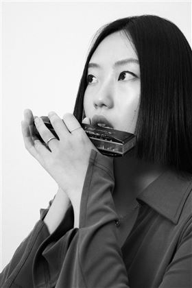 Une femme aux cheveux mi-longs tient une petite boîte dans sa main et regarde en haut. Elle porte un haut gris simple et la prise de vue est en noir et blanc.